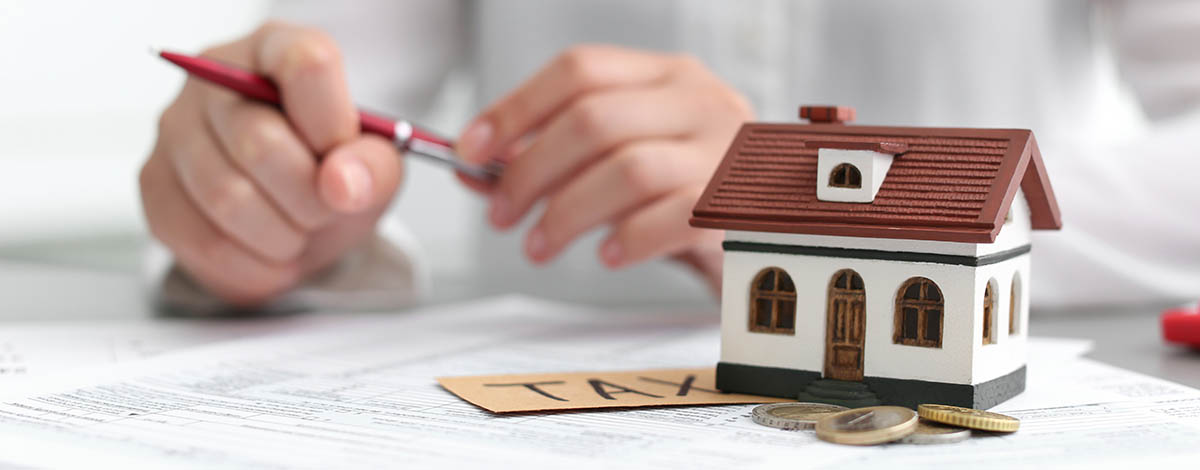 A person holding a pen near a small house model and TAX blocks.
