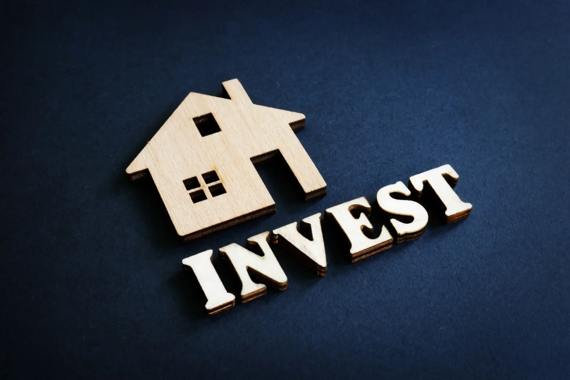 Wooden house model with the word INVEST beside it on a dark background.