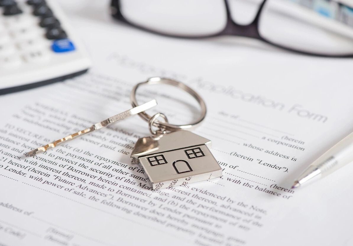 A keychain with a house-shaped pendant rests on a mortgage application form, surrounded by a pair of glasses, a calculator, and a pen.