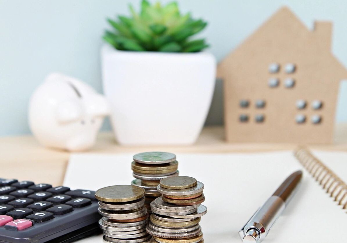 A calculator, stacks of coins, a pen, a notebook, a piggy bank, and a model house sit on a desk next to a potted succulent plant.