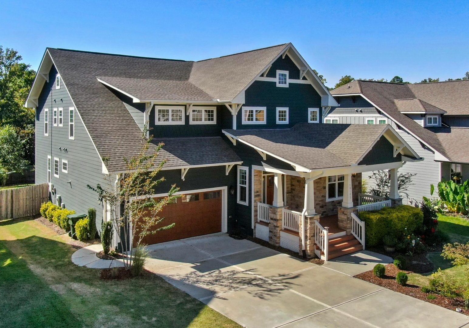 A two-story house with a dark exterior, white trim, and a covered front porch. It has a two-car garage and a driveway, surrounded by a well-maintained lawn and neighboring homes.